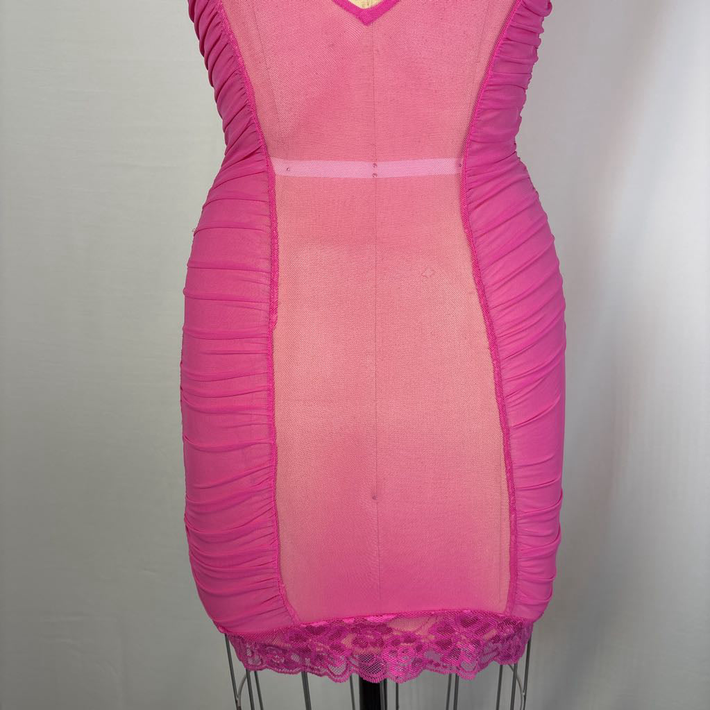 A bright pink bodycon mini dress displayed on a dress form. The dress features ruched side panels, a sheer mesh center panel, a V-neckline, and a ruffled lace hem.