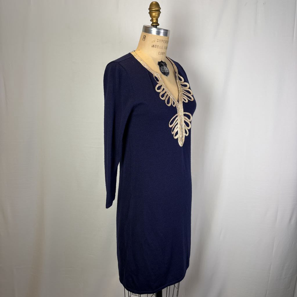 A navy blue knit tunic dress displayed on a vintage wooden dress form mannequin against a white fabric backdrop. The dress features a deep V-neckline embellished with cream-colored lace or ribbon trim.