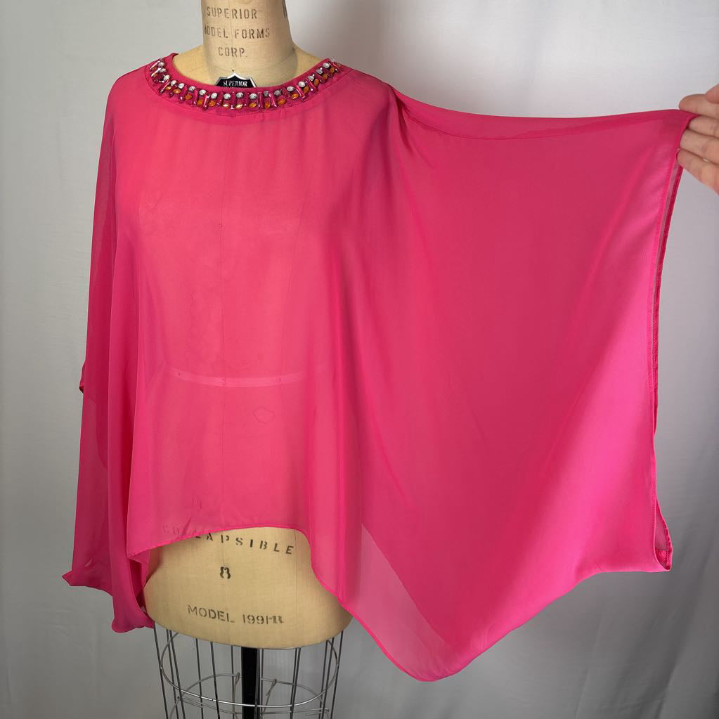 A bright pink sheer poncho-style top displayed on a dress form, with one side held out by a hand. The neckline is embellished with small rhinestone-like beads, and the garment has a loose, flowing silhouette.