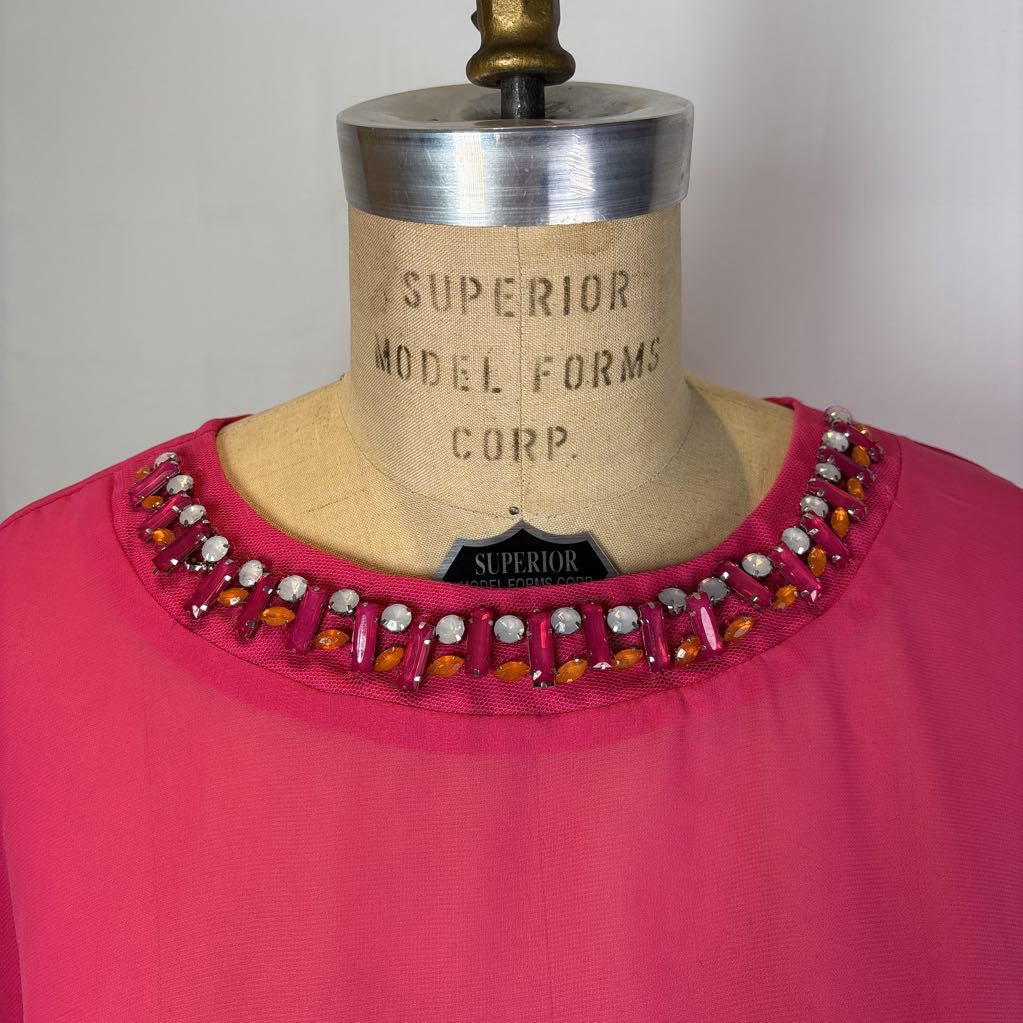 A close-up of a bright pink garment displayed on a vintage dress form mannequin. The neckline is decorated with a row of embellished beads and crystals in pink, orange, and white.