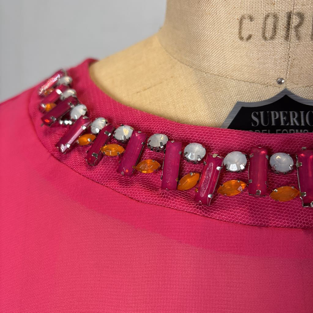 A close-up of a bright pink garment on a beige dress form, featuring a decorative neckline embellished with magenta, orange, and silver rhinestones.