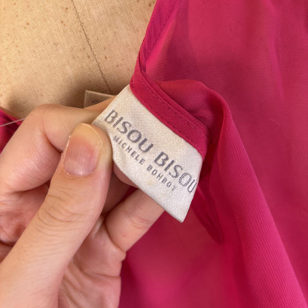 A close-up of a hand holding a white fabric tag that reads 'BISOU BISOU MICHELE BOHMOY' attached to a bright pink garment.