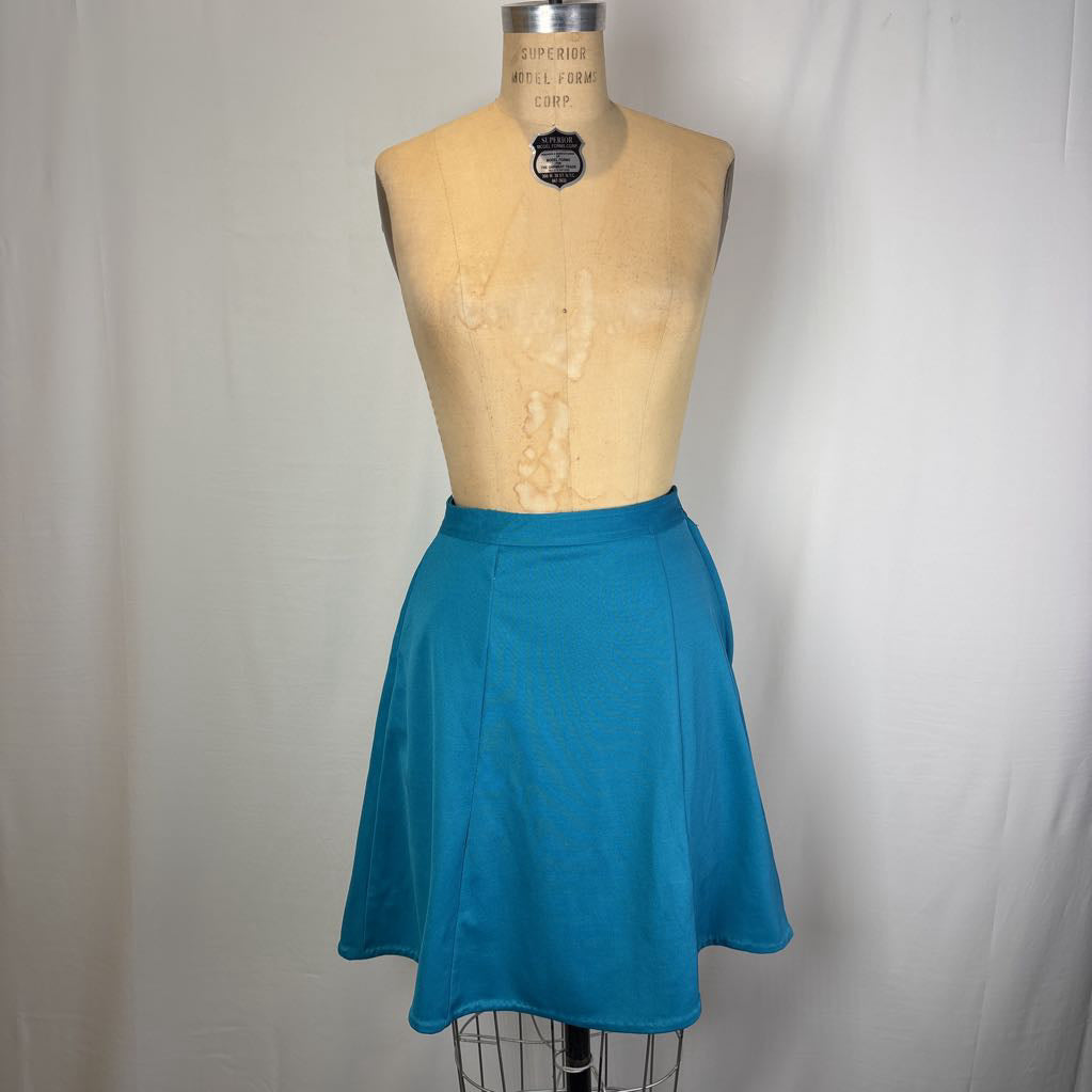 A bright teal blue flared skirt displayed on a vintage tan dress form mannequin against a white fabric backdrop.