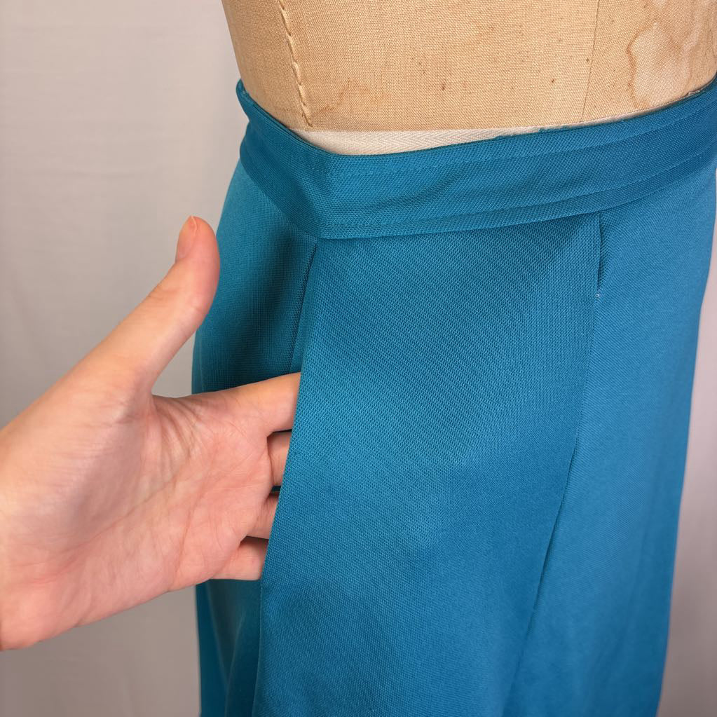 A close-up of teal blue trousers displayed on a beige dress form, with a hand pointing to the side pocket area.