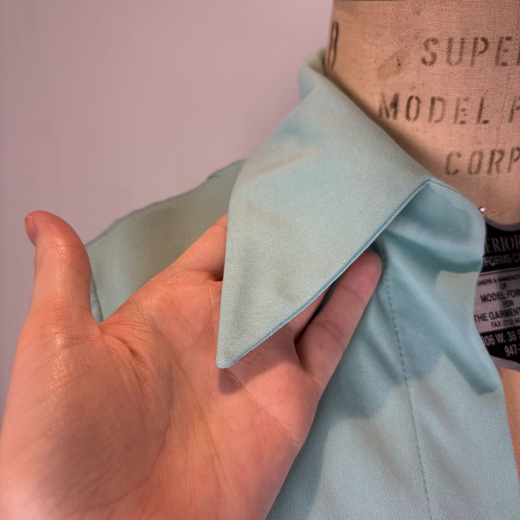 A close-up of a light blue-green collared shirt on a mannequin, with a hand holding the collar to show its shape and fabric texture.