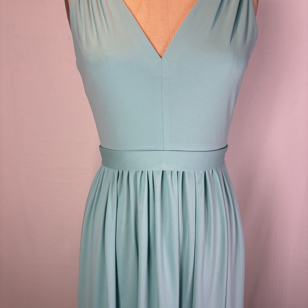 A sleeveless dusty blue-green dress displayed on a mannequin against a soft pink background. The dress features a V-neckline, a matching waistband, and a gathered skirt.