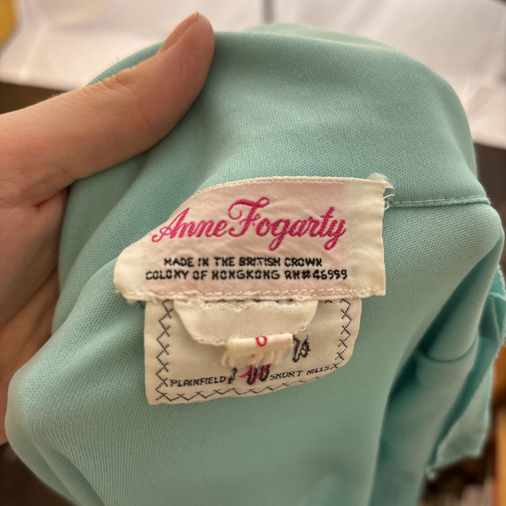 A close-up of a hand holding a mint green garment, showing the brand label 'Anne Fogarty' sewn onto the inside collar.