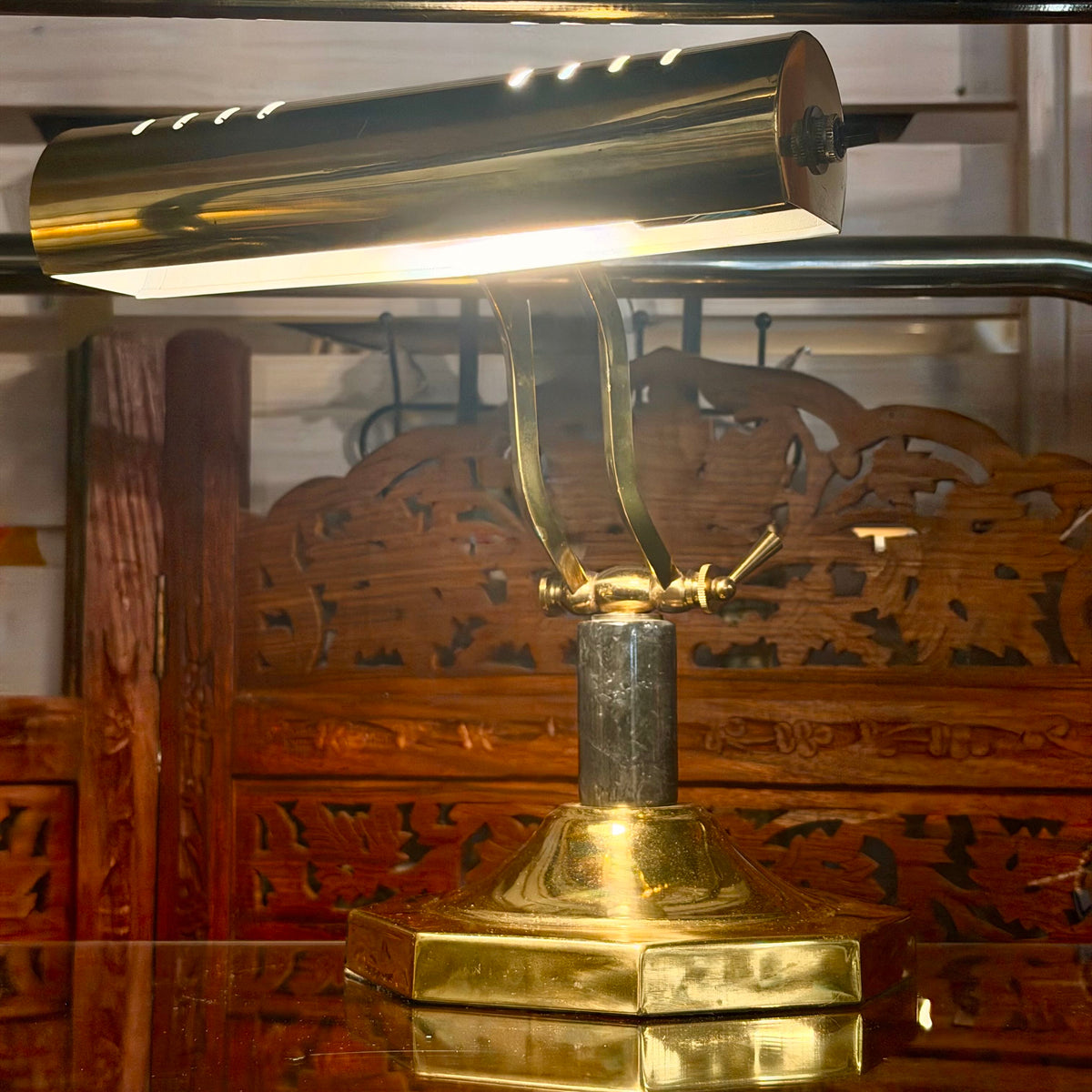 A polished brass desk lamp with a rectangular shade, adjustable curved neck, and hexagonal base, illuminated on a reflective surface with a carved wooden background.