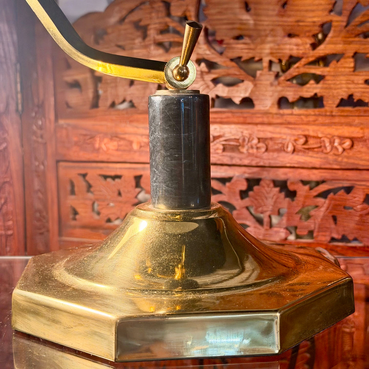 A close-up of a brass desk lamp base with a hexagonal pedestal base, a central cylindrical neck, and a curved arm with a black cord, set against a carved wooden background.