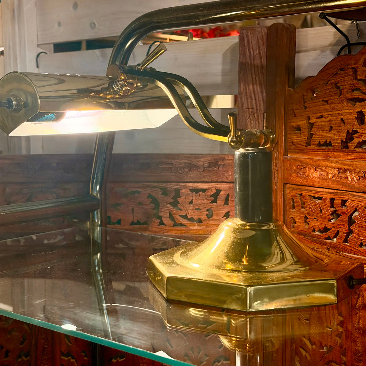 A brass desk lamp with a hexagonal base, adjustable curved arm, and white glass shade, shown lit on a glass shelf in front of carved wooden panels.