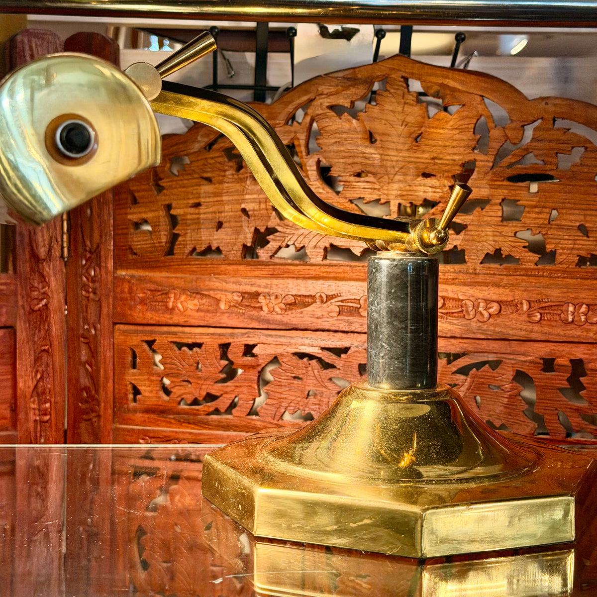 A brass desk lamp with a curved arm and hexagonal base, shown in close-up against a carved wooden background.