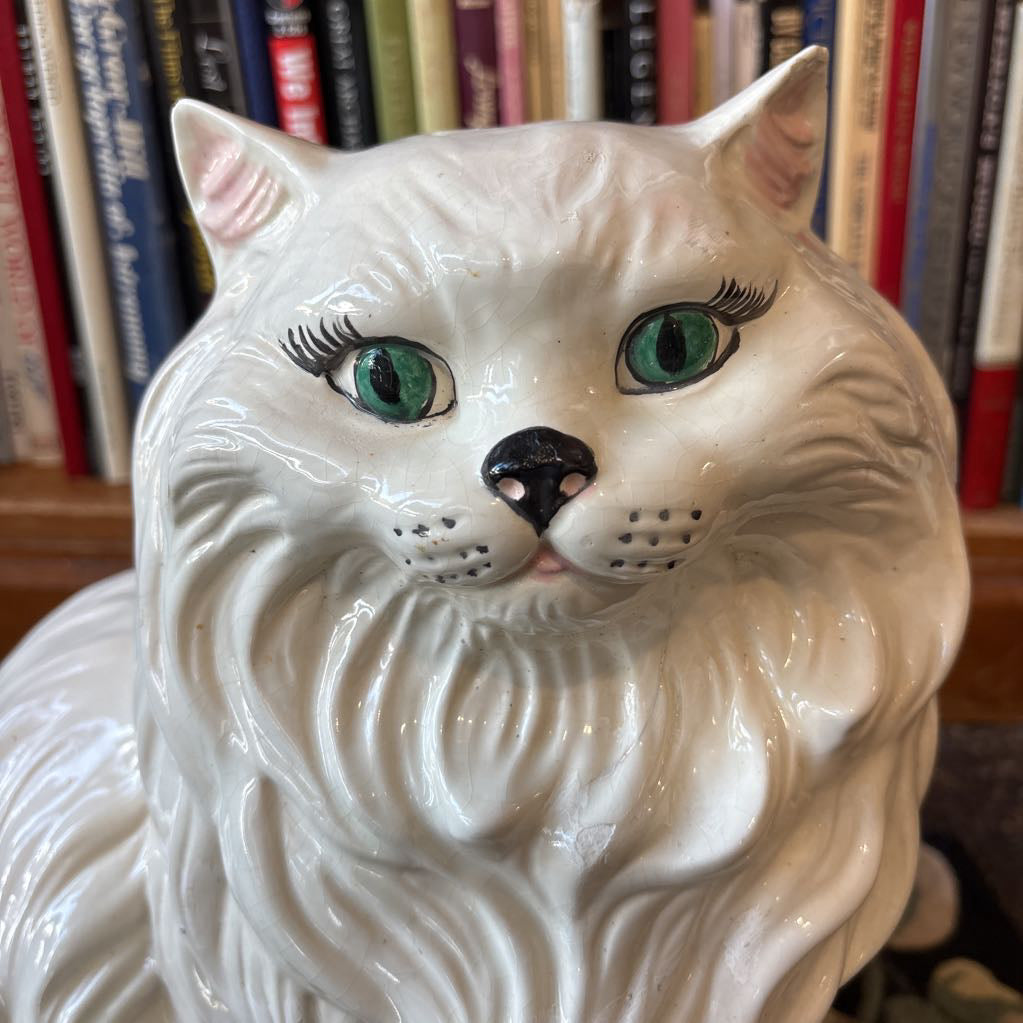 A large white ceramic cat figure with green eyes and long eyelashes sitting on a floral rug against a bookcase.