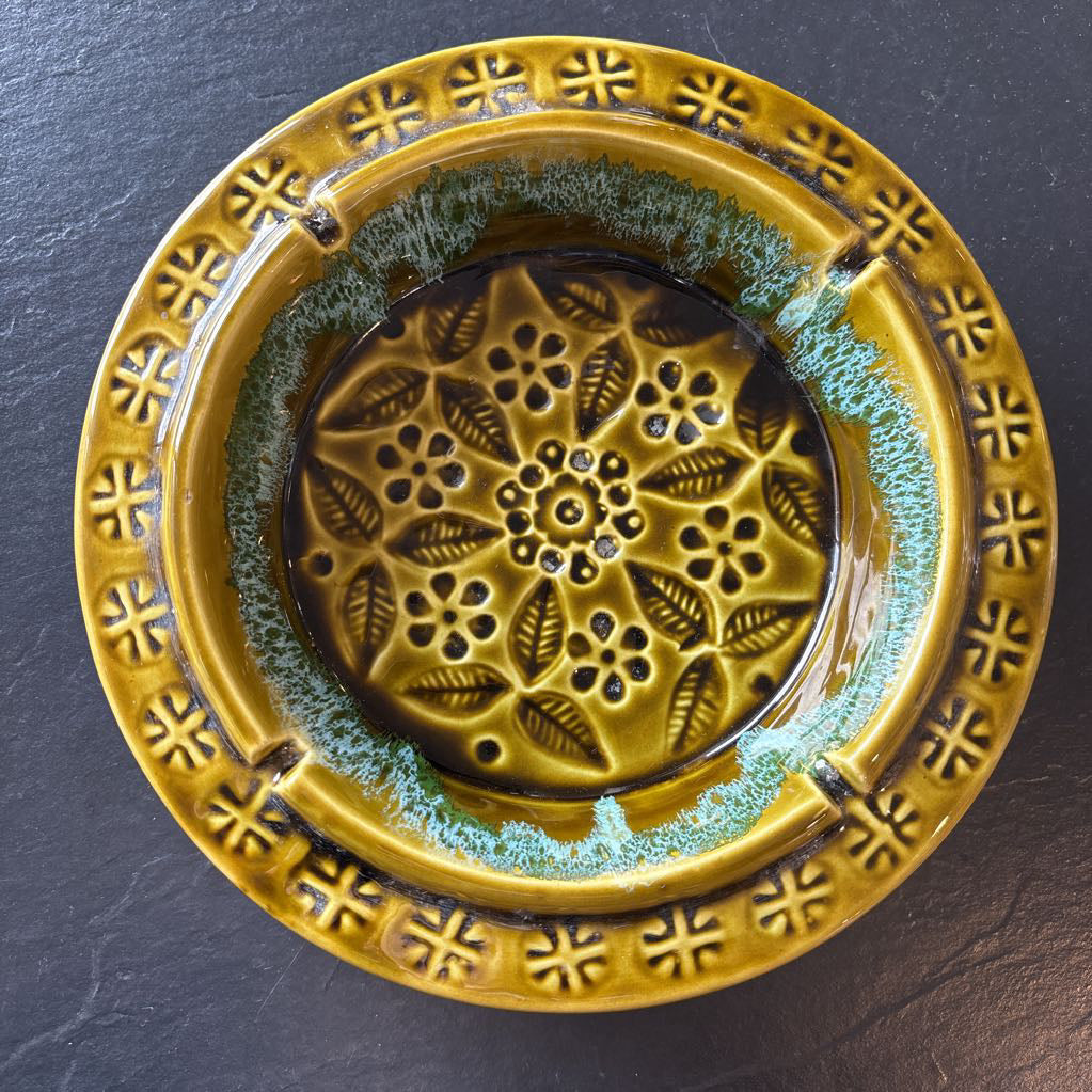 A round ceramic ashtray with a glossy mustard yellow and olive green glaze, featuring a raised floral border and a central leaf pattern, set on a dark slate background.