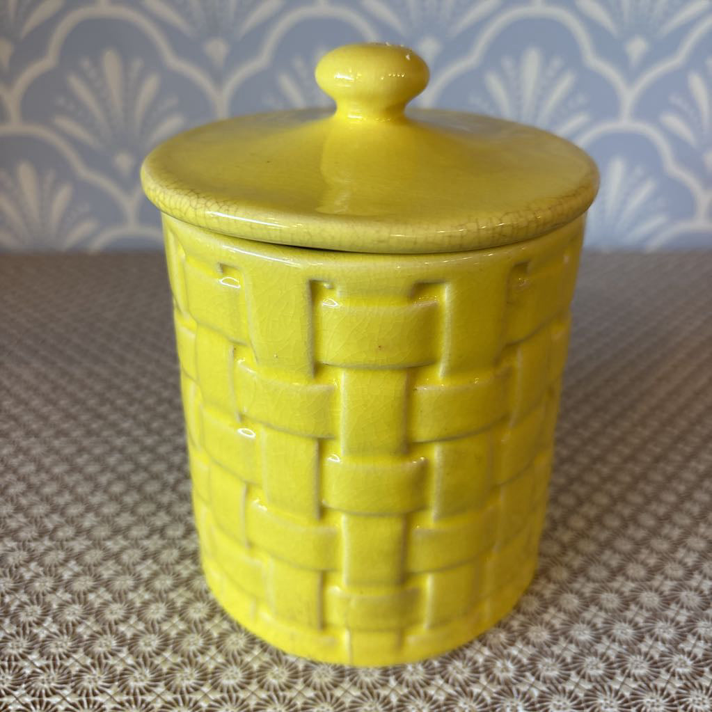 A yellow basket weave textured ceramic canister with lid on a wallpapered surface.