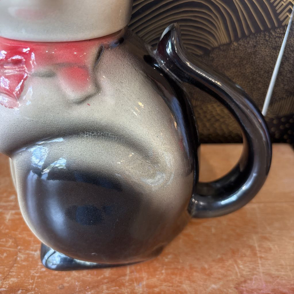 A ceramic teapot of a tuxedo cat with green eyes and a pink bowtie. Its arm is the spout and its tail the handle. It sits on a wooden surface against a black and gold wall.