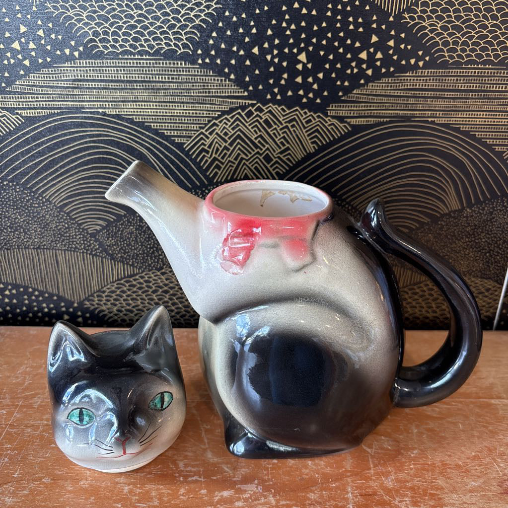 A ceramic teapot of a tuxedo cat with green eyes and a pink bowtie. Its arm is the spout and its tail the handle. It sits on a wooden surface against a black and gold wall.