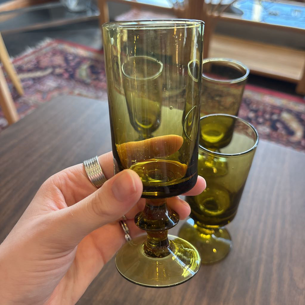 A hand holds a translucent olive-green glass goblet with a flared base, with two more matching goblets blurred in the background on a wooden table.