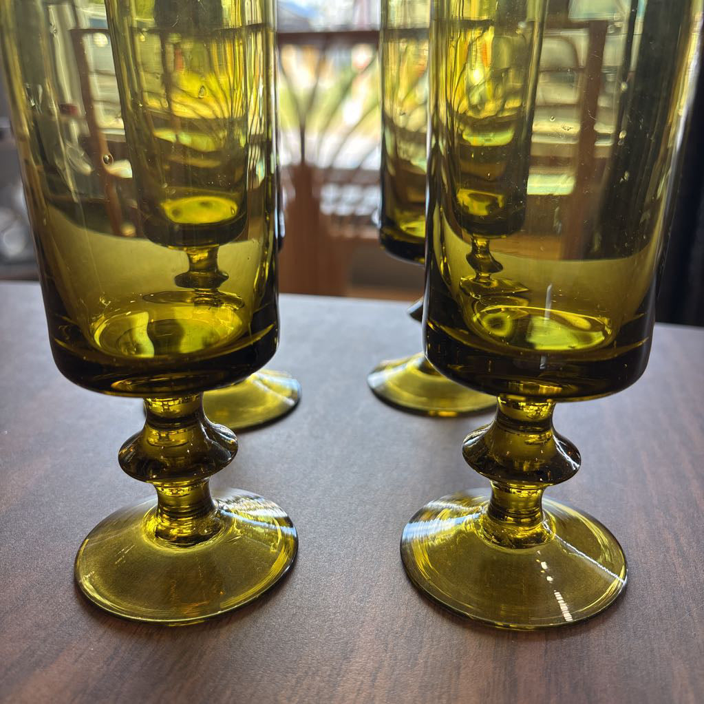 Three translucent olive-green glass goblets with flared bases arranged on a wooden tabletop, with a softly blurred indoor background visible through the glass.