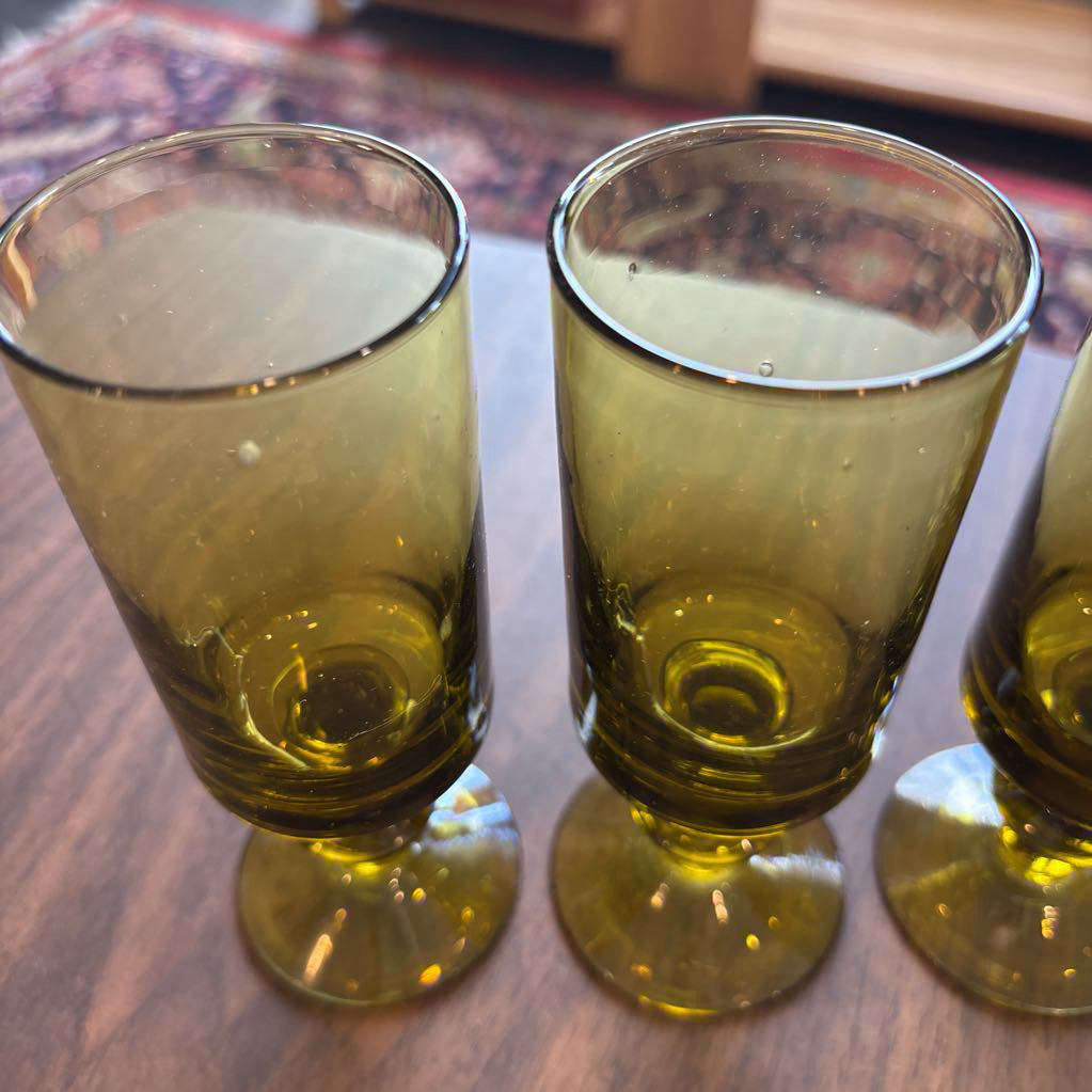 Three vintage olive-green glass goblets with stems and bases, arranged on a wooden tabletop.