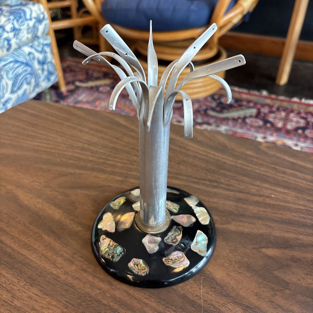 A silver metal jewelry display stand shaped like a stylized tree, featuring a textured central column and multiple curved leaf-like branches, mounted on a round black base decorated with iridescent shell pieces, shown on a wooden table.