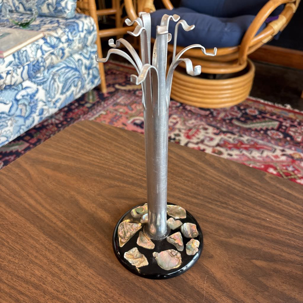 A silver metal jewelry stand designed like a tree trunk with multiple curved branches, standing on a black circular base decorated with small polished stones, placed on a wooden table in a living room.