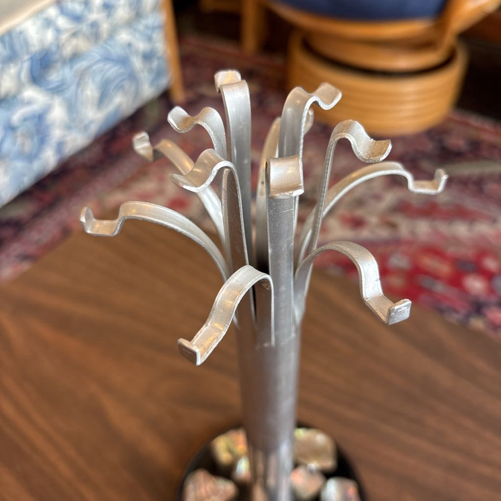 A silver metal jewelry stand with multiple curved hooks, shown on a wooden table with a blurred home background.