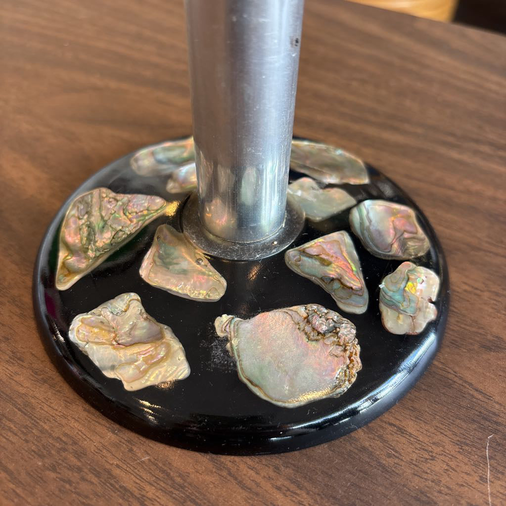 A round black glass or resin stand with multiple iridescent abalone shell pieces around a central silver metal pole, shown on a wooden tabletop.