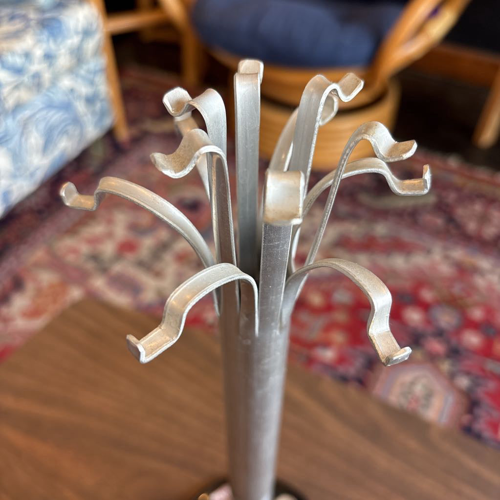 A silver metal jewelry stand with multiple curved hooks, shown standing on a wooden table in a furnished room with a patterned rug and chairs in the background.