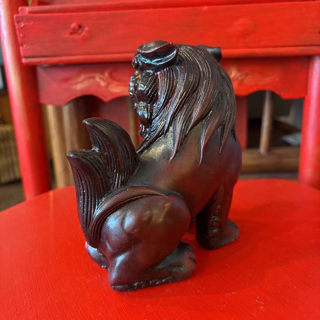 A dark brown foo dog figurine with a muscular build and detailed mane, positioned on a red surface with a red wooden chair in the background.