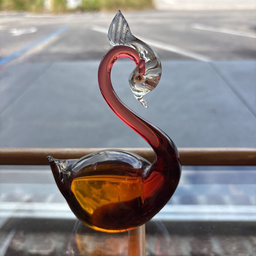 A decorative glass swan figurine with a curved neck, featuring amber-brown and translucent red glass on the body and head, and a clear glass beak, displayed on a reflective surface with a blurred outdoor background.