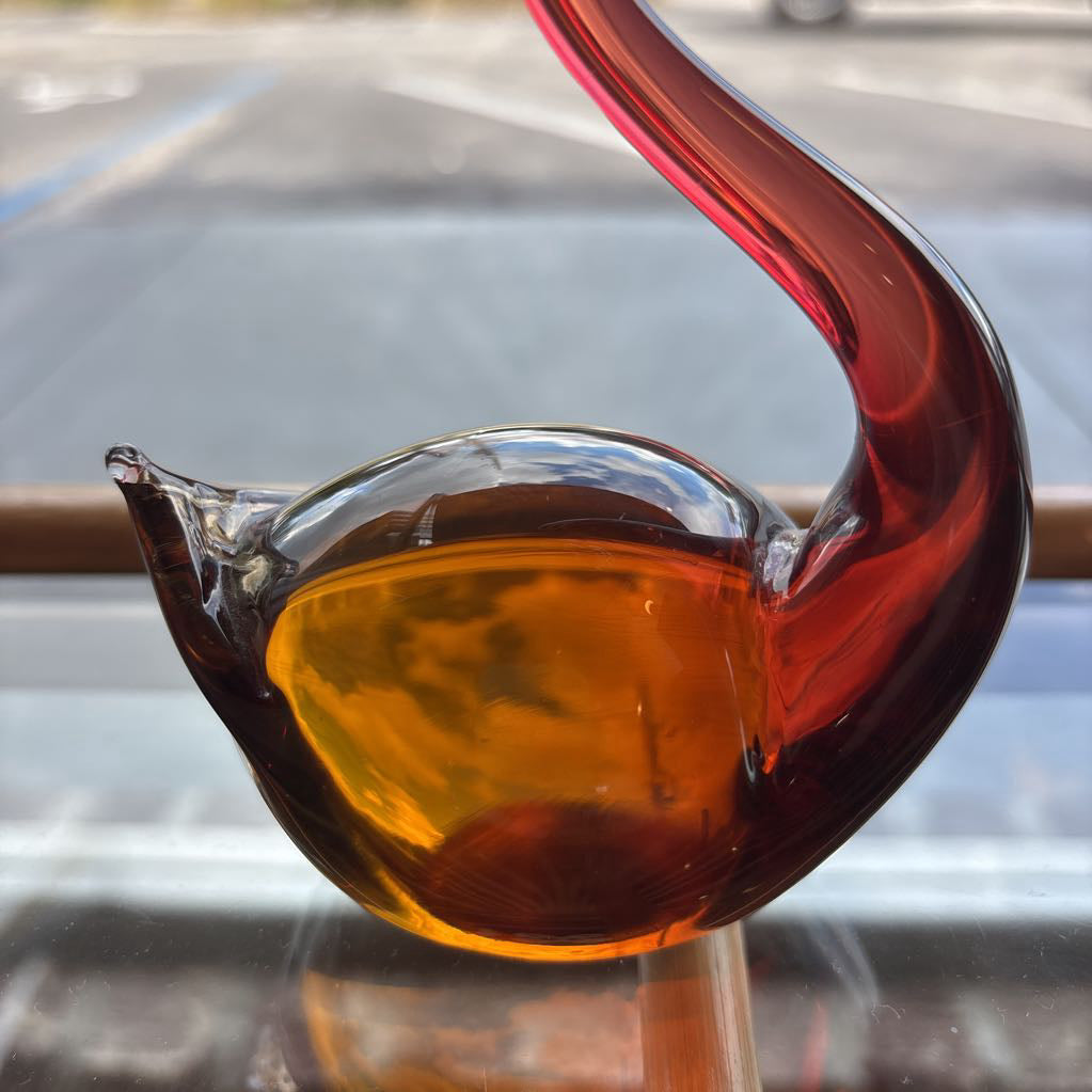 A sculptural glass bird figurine with a curved neck and spout, featuring a translucent amber-yellow body and a deep red neck, photographed on a reflective surface with a blurred outdoor background.