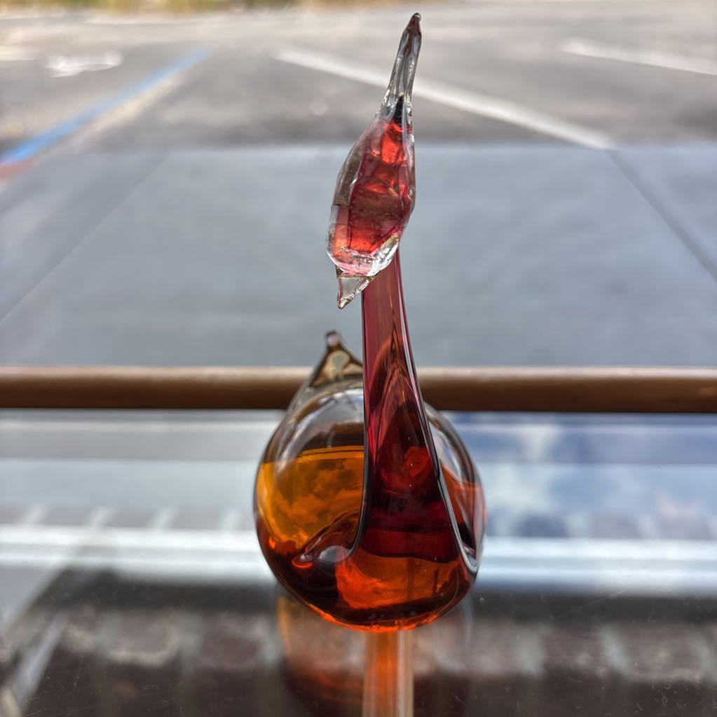 A hand-blown glass swan figurine with a glossy amber and deep red finish, positioned on a reflective glass surface with a blurred outdoor parking lot background.