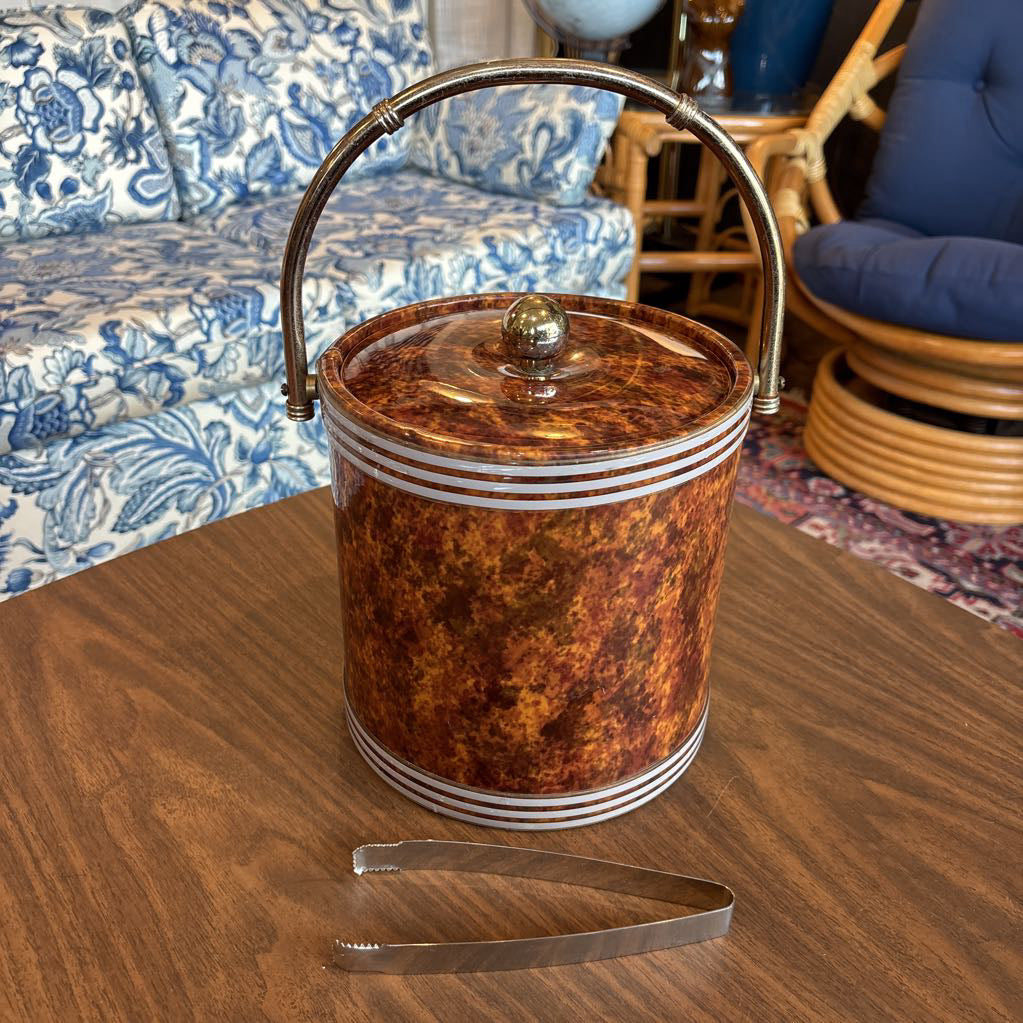 A brown speckled ice bucket with white horizontal stripes and the top and bottom and pair of tongs in a room setting.