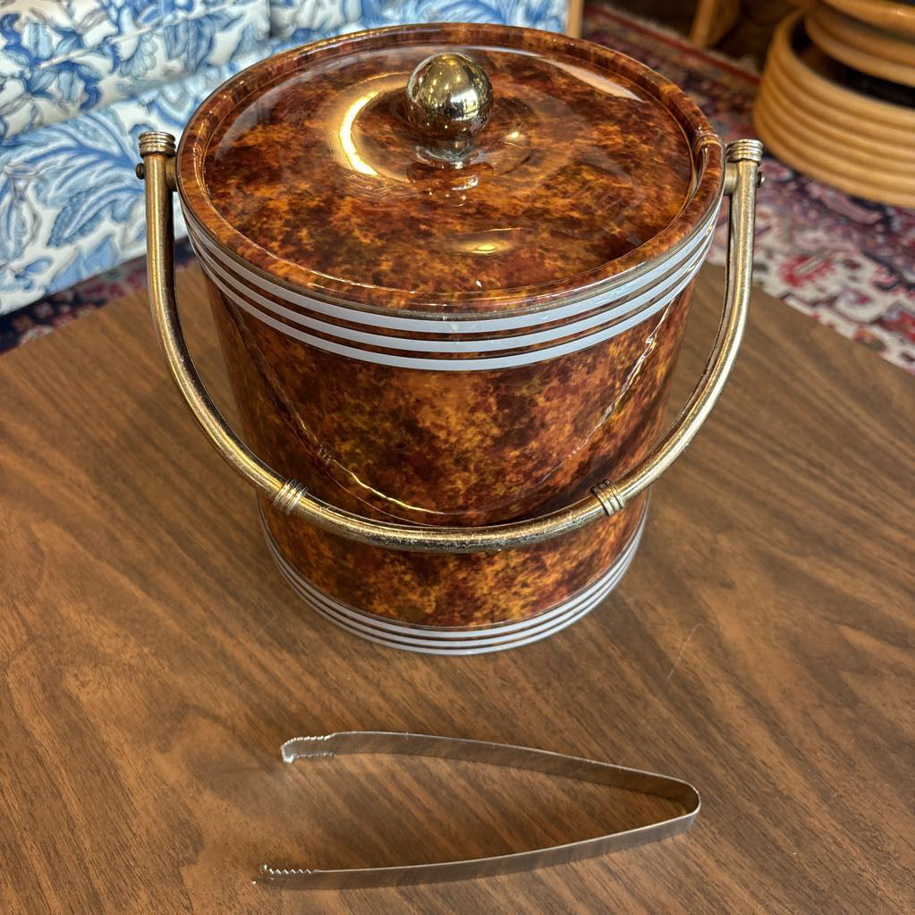 A brown speckled ice bucket with white horizontal stripes and the top and bottom and pair of tongs in a room setting.