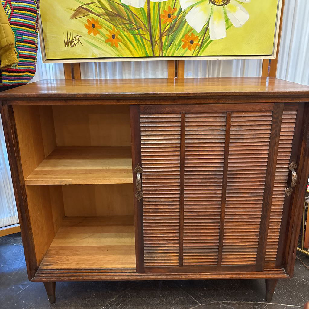 A mid-century modern wooden sideboard cabinet with a warm medium-brown finish, featuring two louvered doors on the right and an open two-shelf compartment on the left. The cabinet is photographed indoors against a white paneled wall, with a colorful floral painting hanging above it.