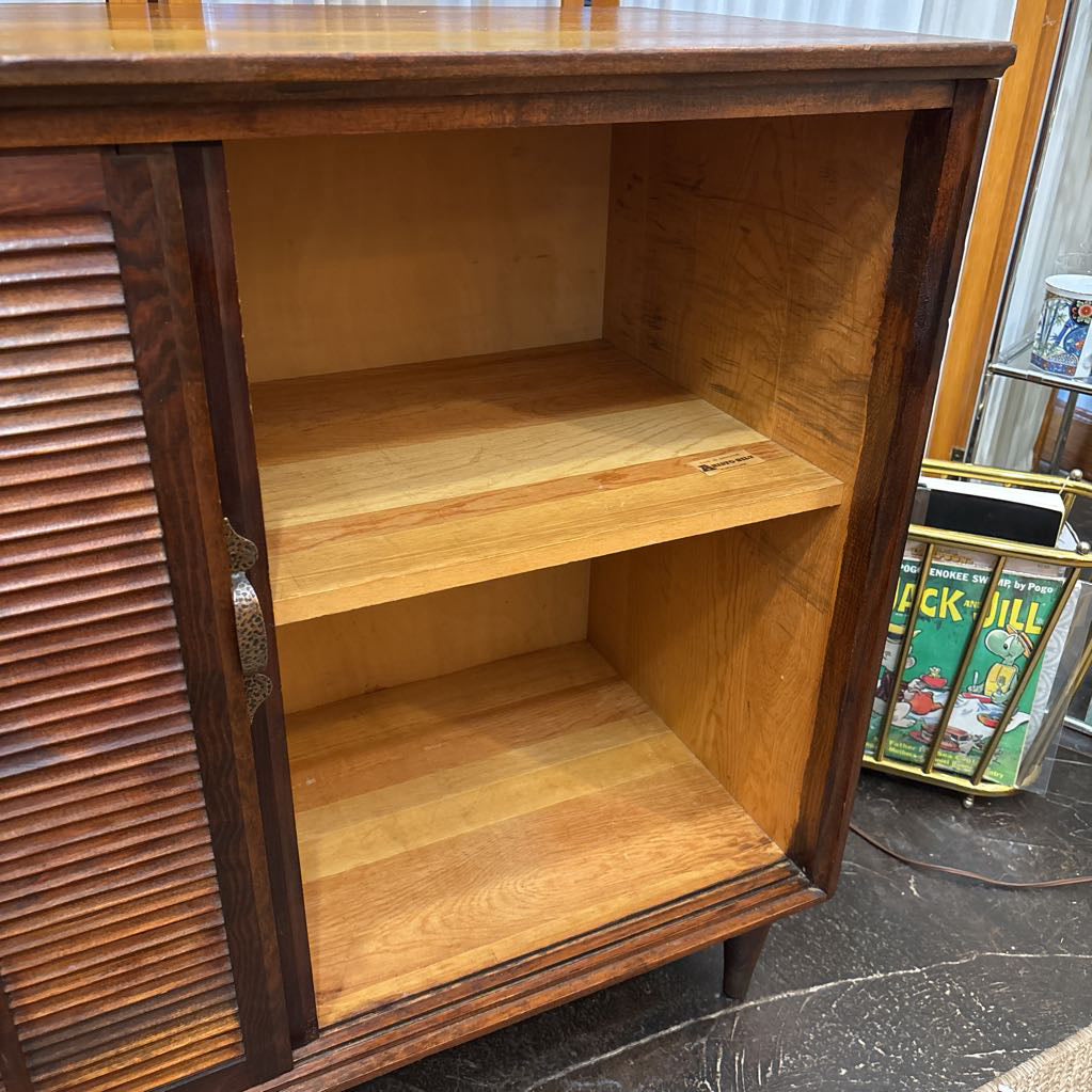 A mid-century modern wooden sideboard cabinet with a dark brown louvered door, open to reveal two light wood shelves, photographed in what appears to be a showroom or home.