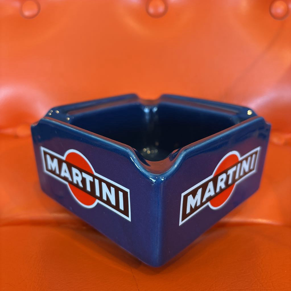 A blue ceramic Martini ashtray featuring the classic red and white logo, set against a bright orange textured background.