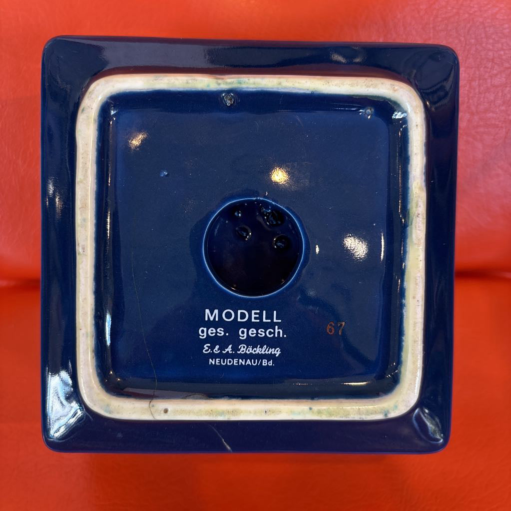 Top-down view of a square dark blue ceramic container or vase base with a raised rim, glossy finish, and printed text reading 'MODELL ges. gesch.