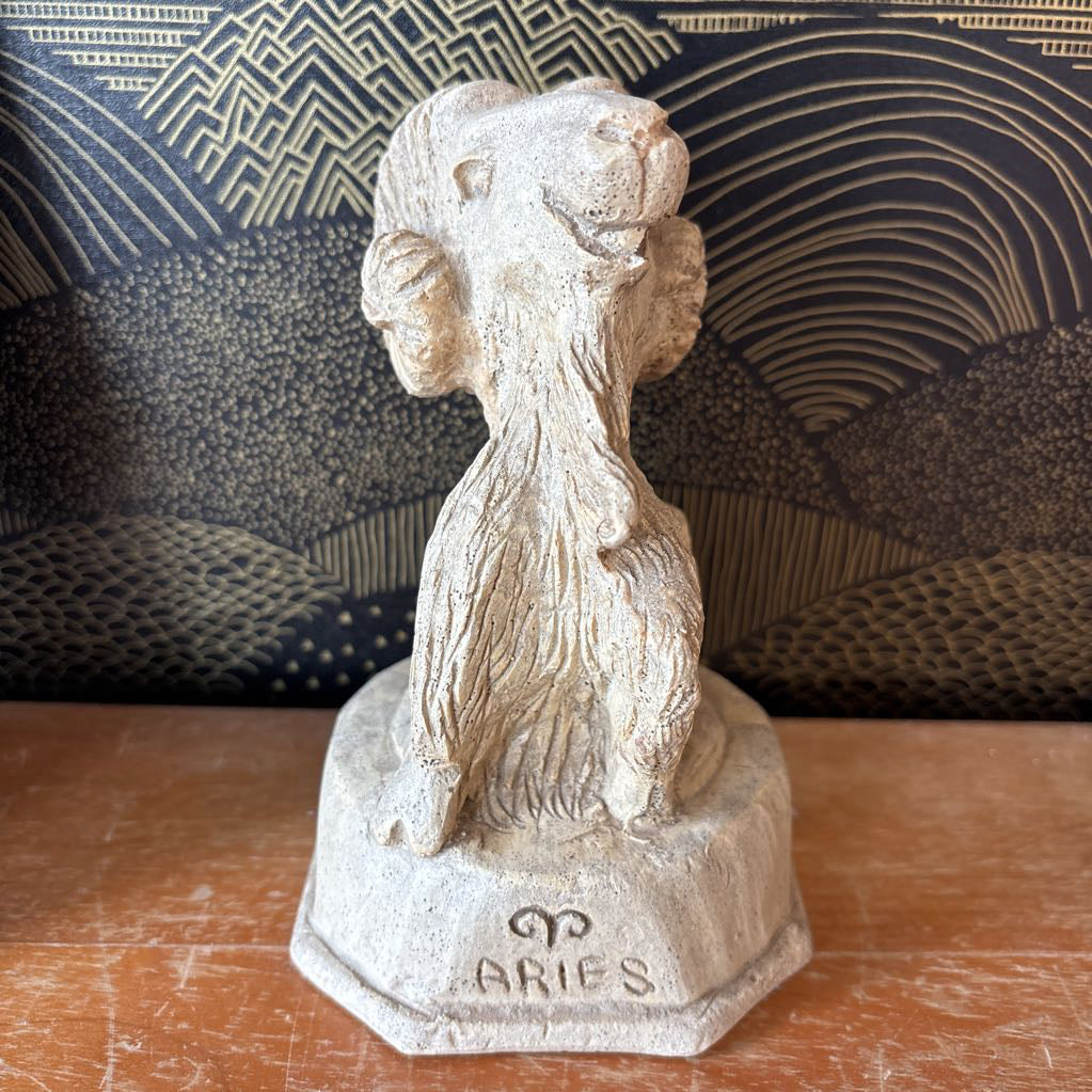 A carved stone figure of a ram on a pedestal. It sits on a wooden surface and a black and gold wallpaper backdrop.