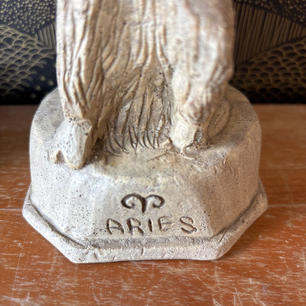 A carved stone figure of a ram on a pedestal. It sits on a wooden surface and a black and gold wallpaper backdrop.