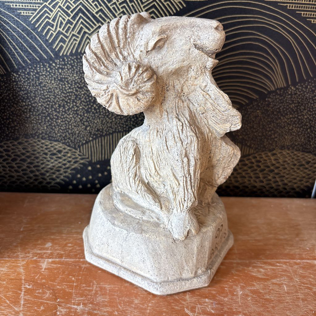 A light beige stone ram bust sculpture on a hexagonal base, displayed on a wooden surface in front of a patterned black and gold chair.