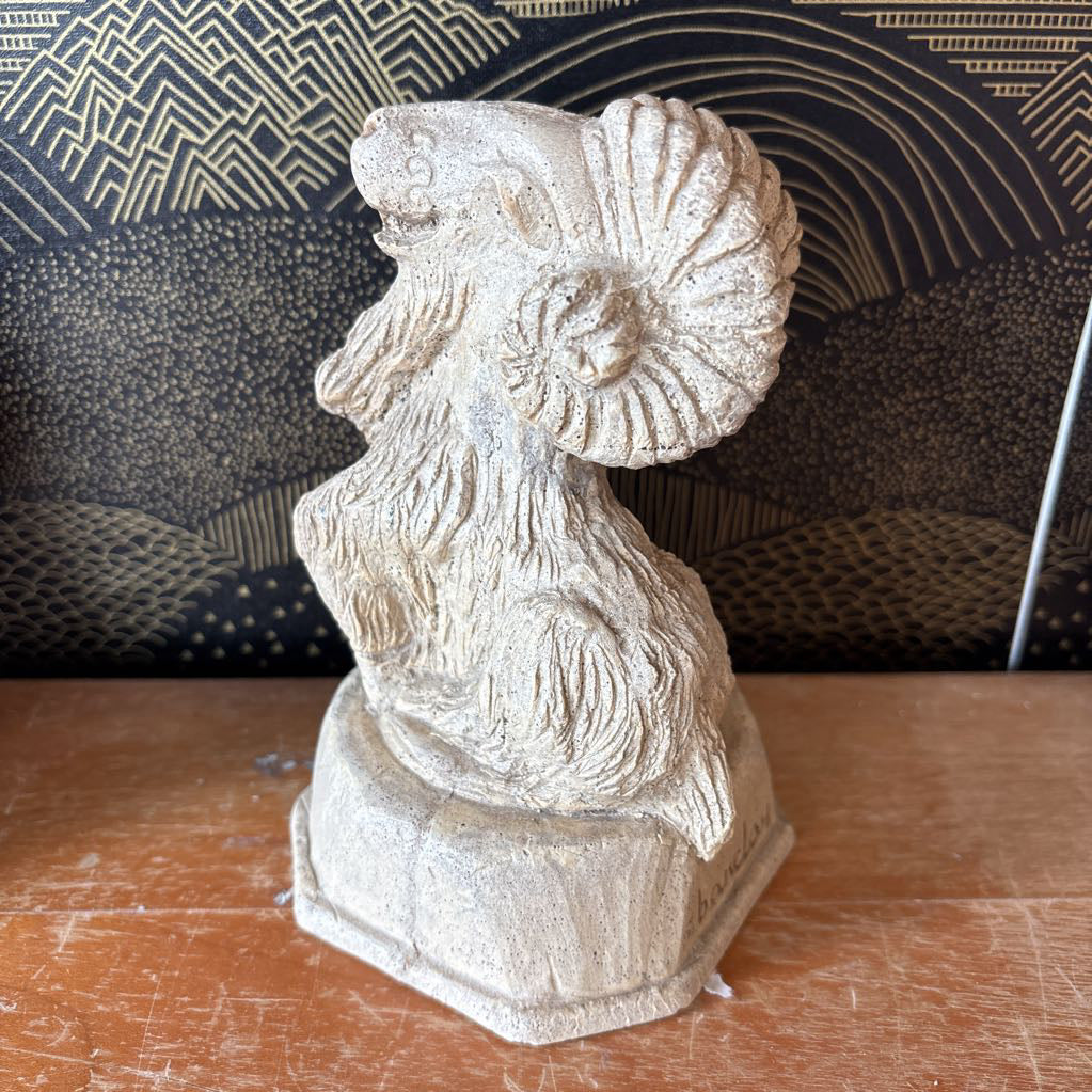A carved stone figure of a ram on a pedestal. It sits on a wooden surface and a black and gold wallpaper backdrop.