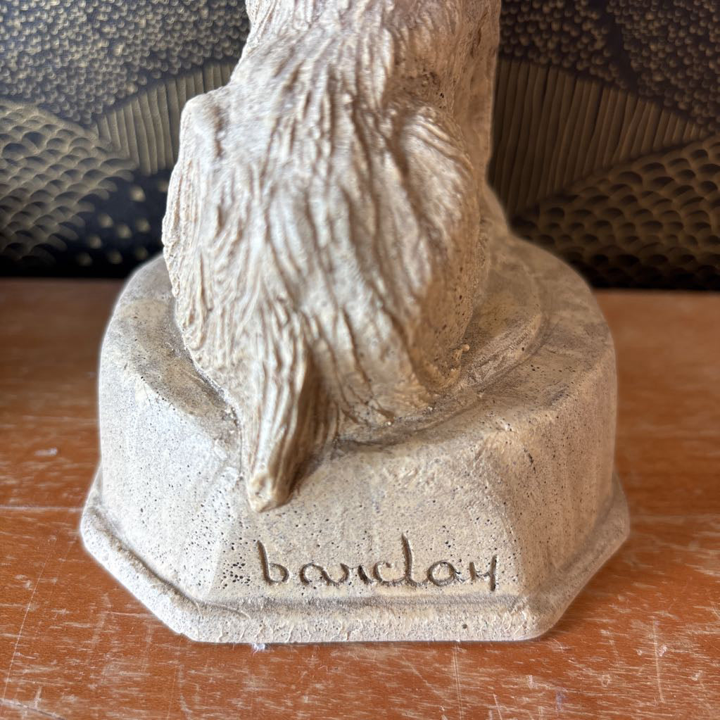 Close-up of a textured stone or concrete figurine base with the name 'barclay' engraved on it, sitting on a wooden surface.