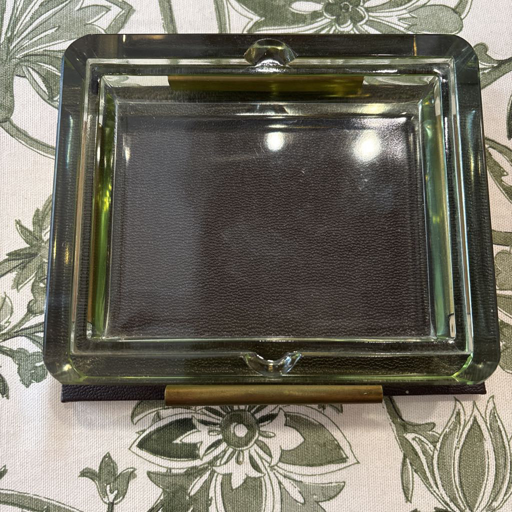 A square glass ashtray with a dark brown textured center and brass-colored metal accents, shown on a green and white floral patterned fabric surface.