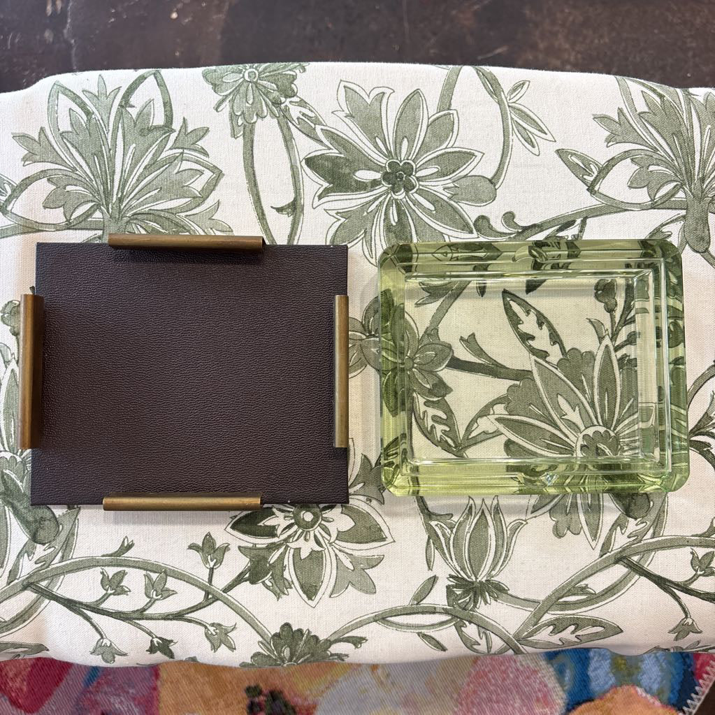 A dark brown leather rectangular tray with brass corner accents sits next to a translucent light green square glass ashtray on a white fabric surface with a green floral print.