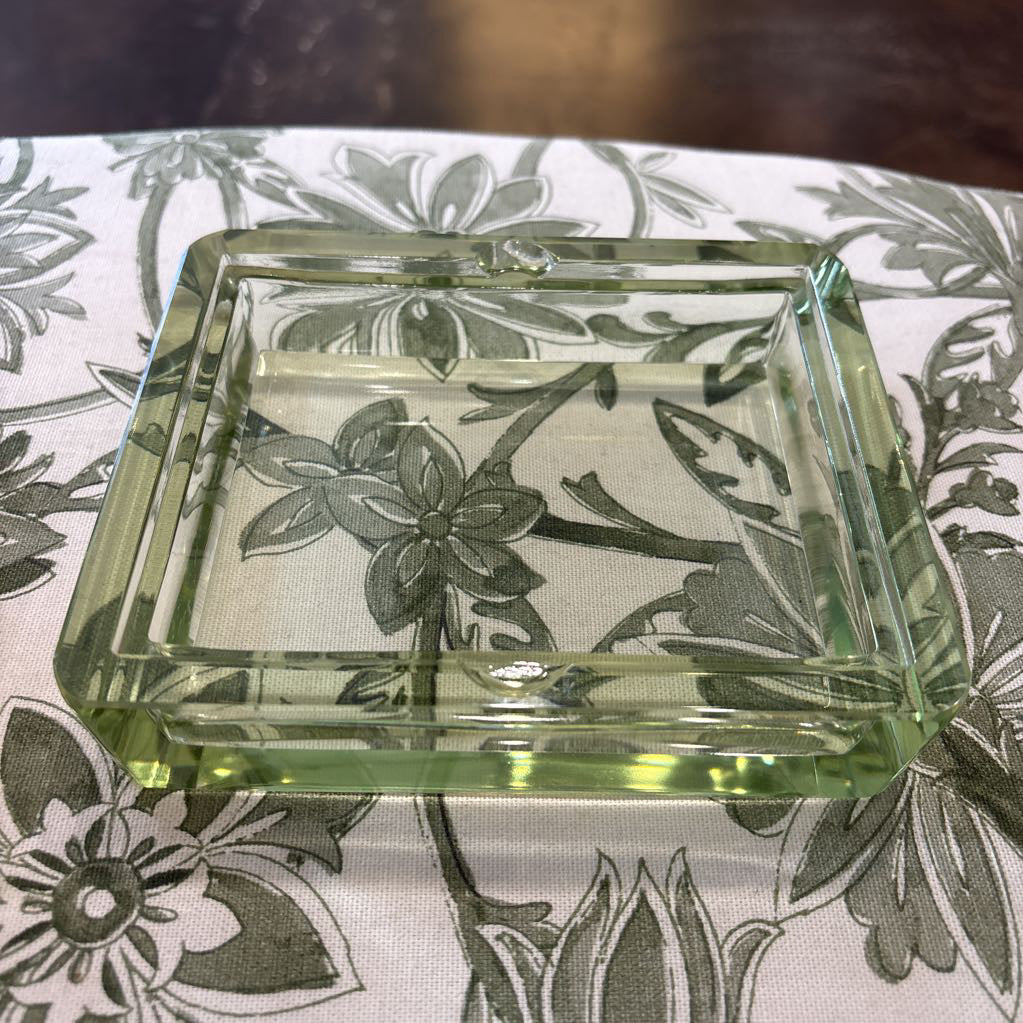 A square clear glass ashtray with a light green tint and embossed floral pattern, resting on a white fabric surface with a gray floral print.