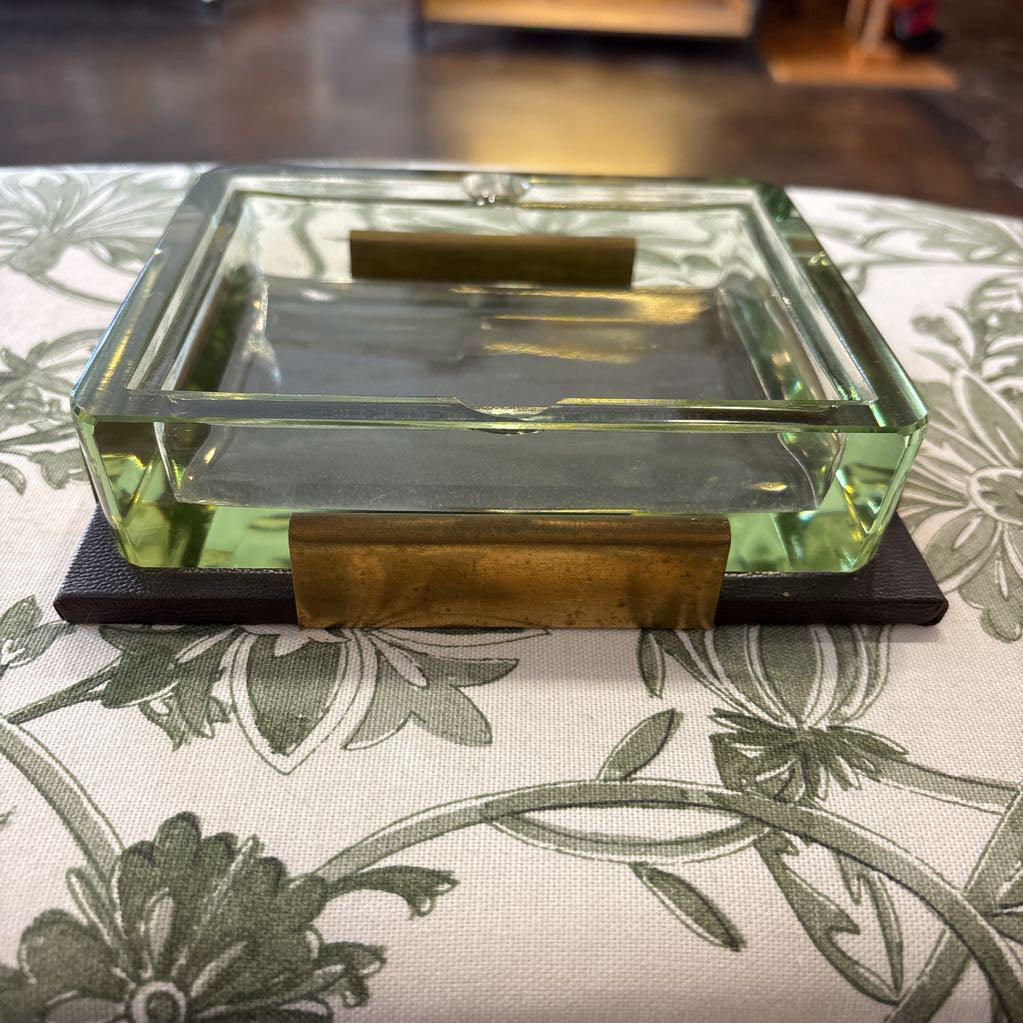A square clear glass tray with a light green tint and brass metal accents, resting on a dark base over a green floral patterned tablecloth.