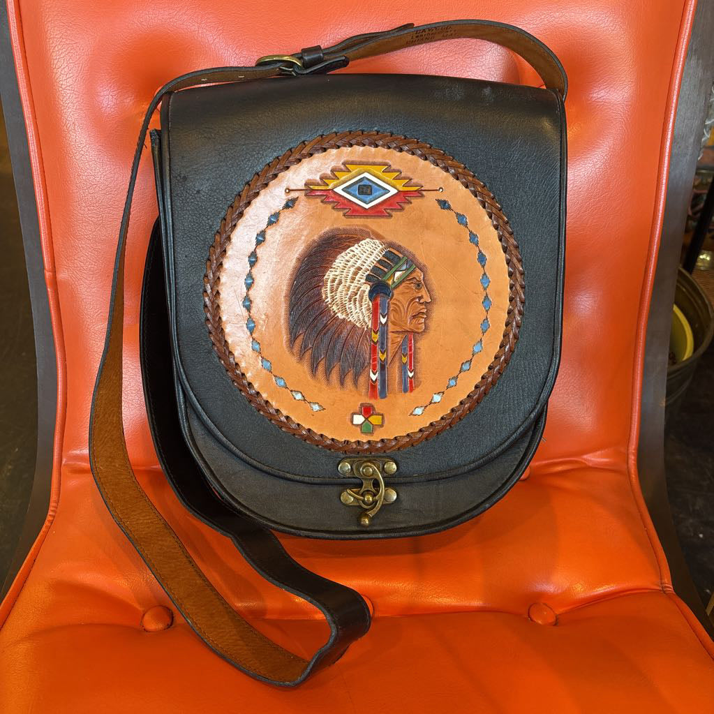 A black leather belt bag with a Native American figure's profile embossed in a circular design on the front. This bag sits on an orange cushion in a room setting.