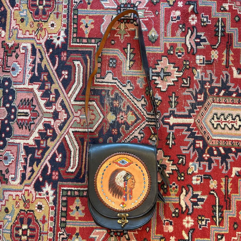 A black leather belt bag with a Native American figure's profile embossed in a circular design on the front. This bag sits on a decorative rug.