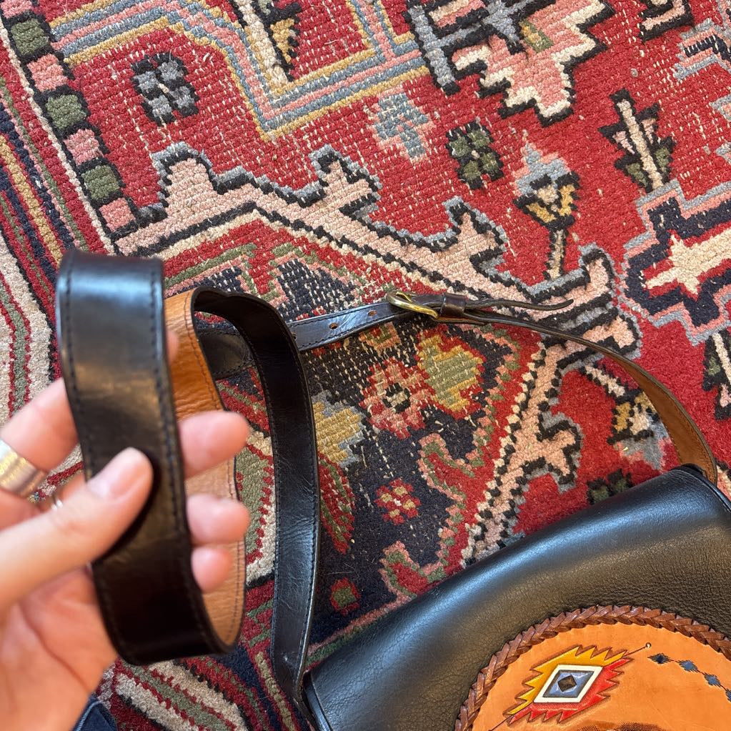 A black leather belt bag with a Native American figure's profile embossed in a circular design on the front. This bag sits on a decorative rug.